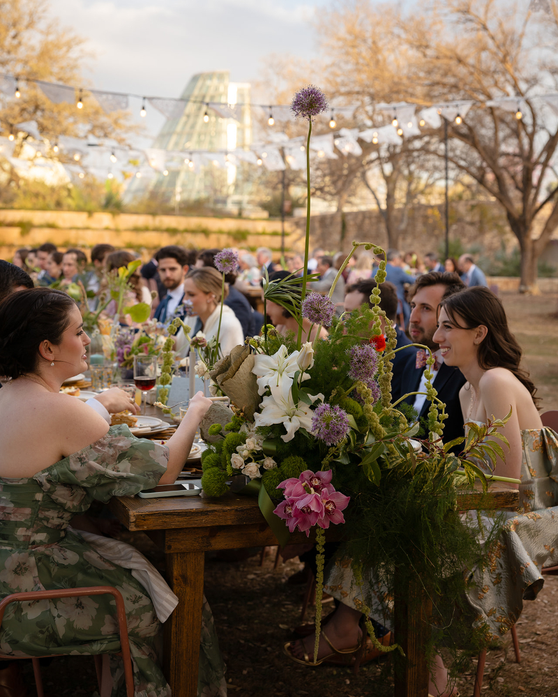 People sit at a long, outdoor table decorated with large floral arrangements, dining and conversing under string lights in a garden setting.