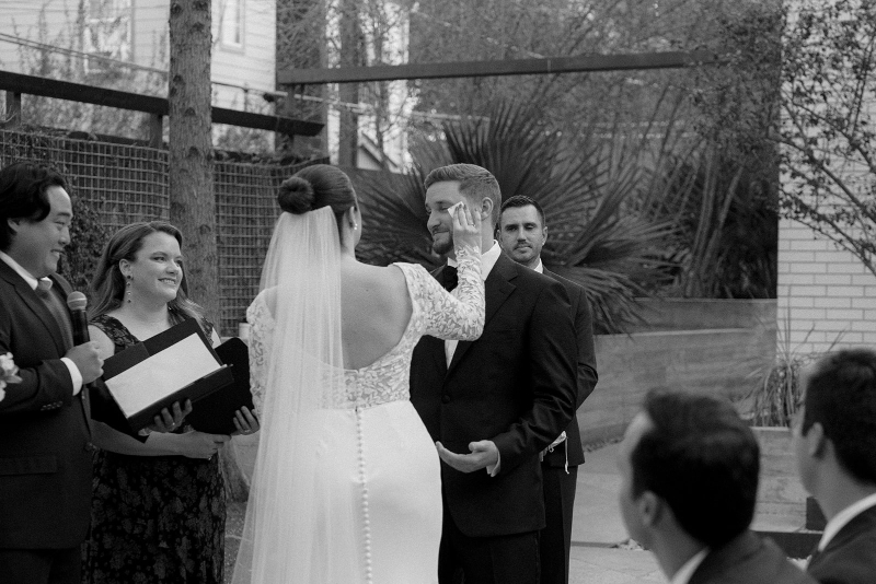 A bride in a long-sleeved dress touches the groom’s face during an outdoor wedding ceremony, with officiants and guests looking on.