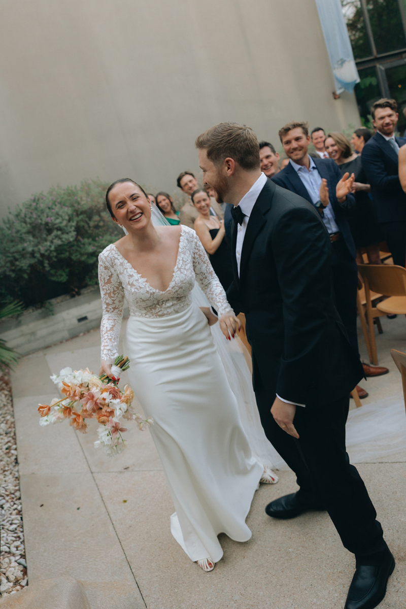 A bride in a white dress and groom in a black tuxedo walk down the aisle smiling, surrounded by applauding guests at an indoor-outdoor wedding ceremony.