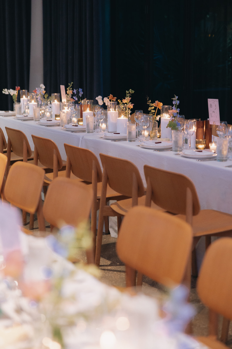 A long dining table set with white tablecloth, glassware, candles, and small floral arrangements, with wooden chairs arranged along both sides, wedding design ideas executed by In Ink Weddings.