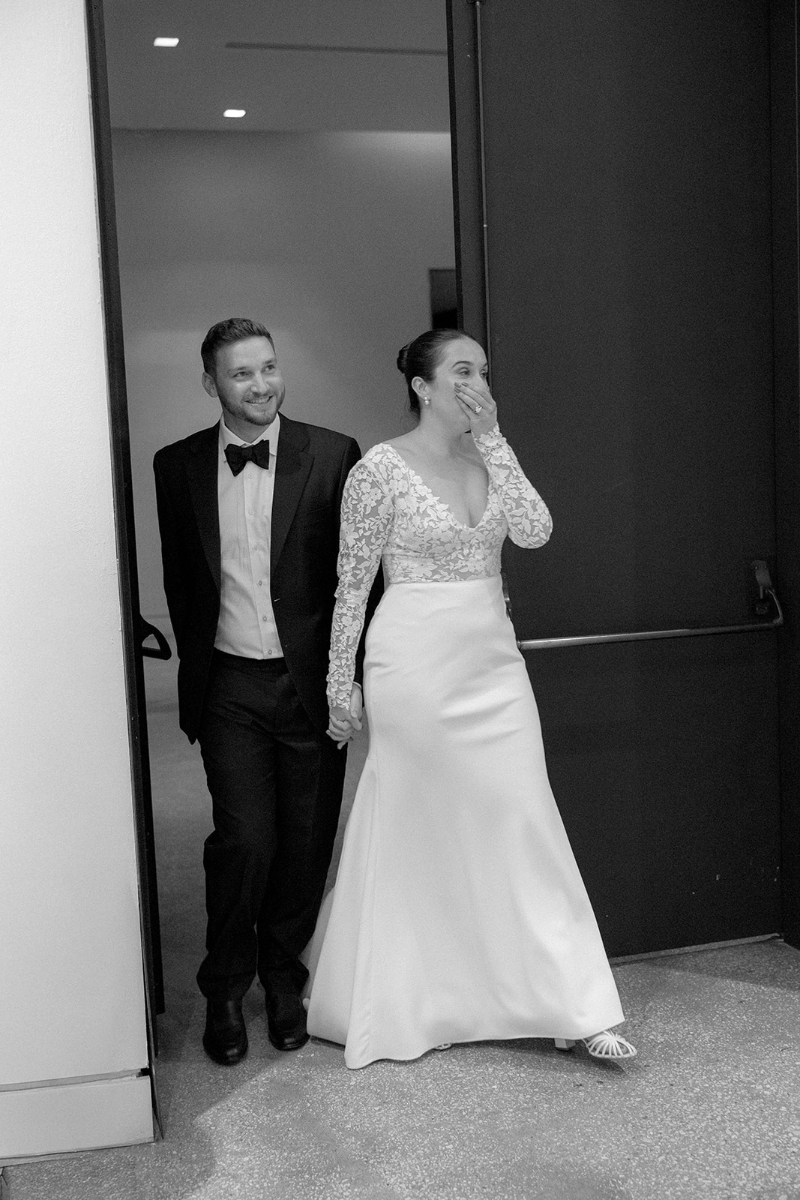 A couple dressed formally, with the woman in a wedding gown and the man in a suit, hold hands and walk through a doorway; the woman covers her mouth with her hand.