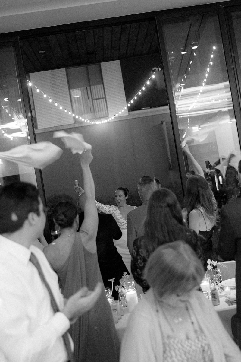 A group of people at an indoor event wave napkins in the air while a woman in a white dress stands smiling near large windows with string lights reflected on the glass.