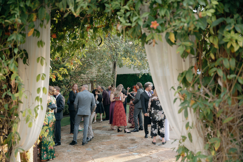 A group of people dressed in formal attire gather and converse outdoors on a stone pathway framed by greenery and white drapes.
