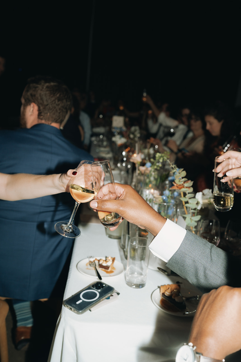 People sitting at a table raise glasses in a toast; the table is set with plates, glasses, flowers, and a smartphone.