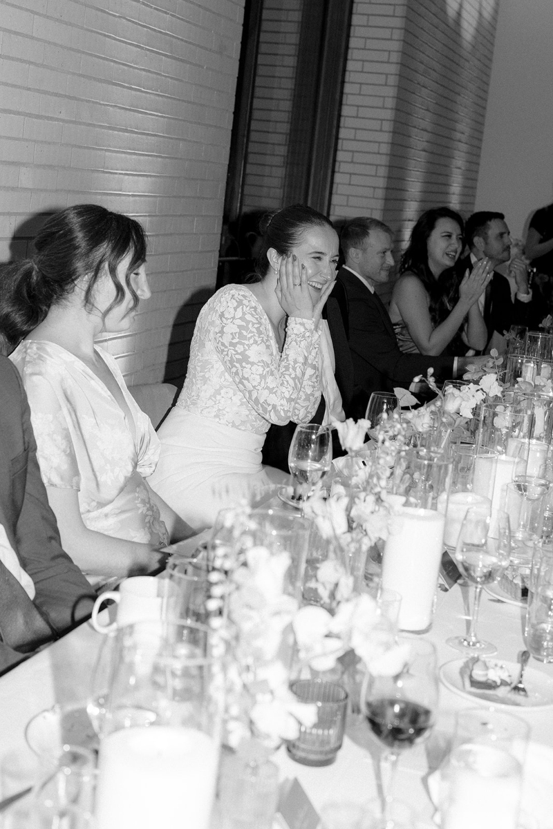 A group of people dressed formally sit at a long table set with glasses, candles, and flowers. Several are smiling and clapping, while one woman in a white dress holds her face, appearing delighted.
