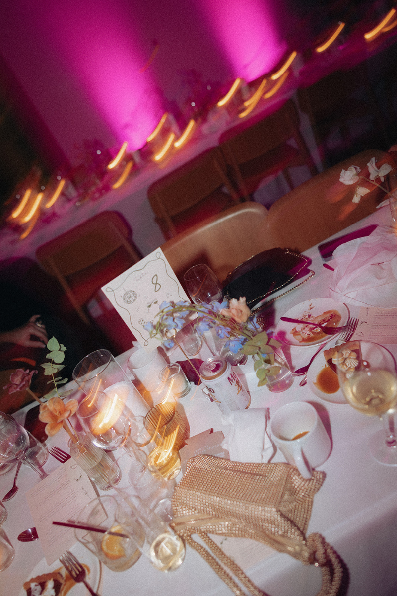 Wedding design ideas for messy luxury weddings including decorated dinner table with empty glasses, candles, plates with leftover food, a menu card, flowers, and a beaded purse, with pink-lit walls and chairs in the background.