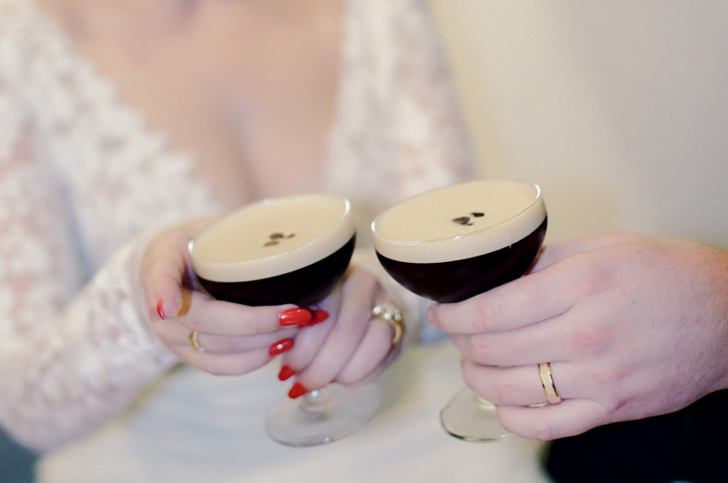 Two people holding espresso martini cocktails in coupe glasses, both wearing wedding bands; one person is in a white lace dress with red nail polish.