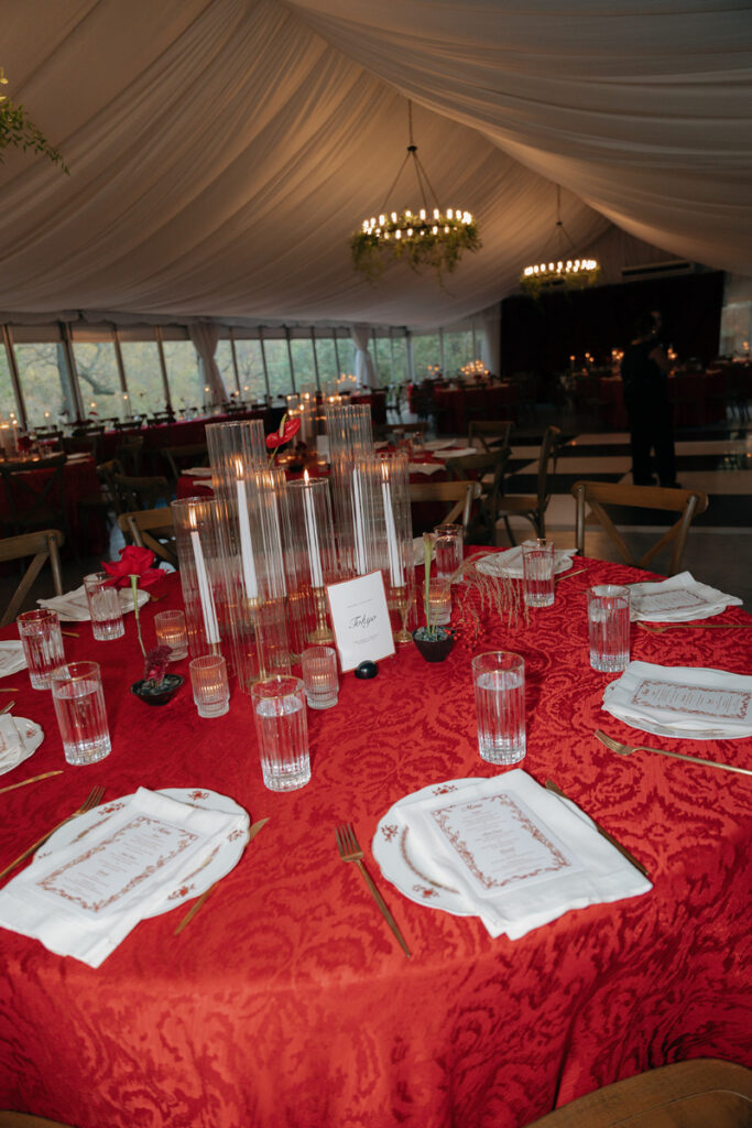 A round table with a red tablecloth is set with plates, glasses, menus, white napkins, and tall glass candle holders in an elegant event tent.