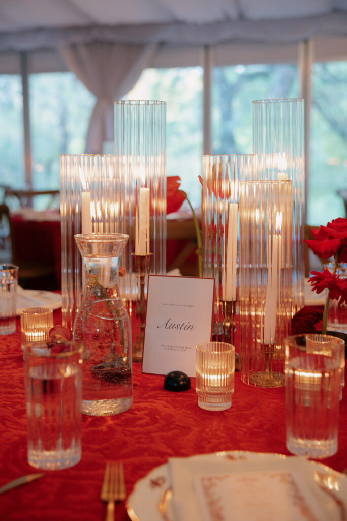 A table set with water glasses, a carafe, gold utensils, a menu card, red flowers, and several lit candles in tall glass holders on a red tablecloth.