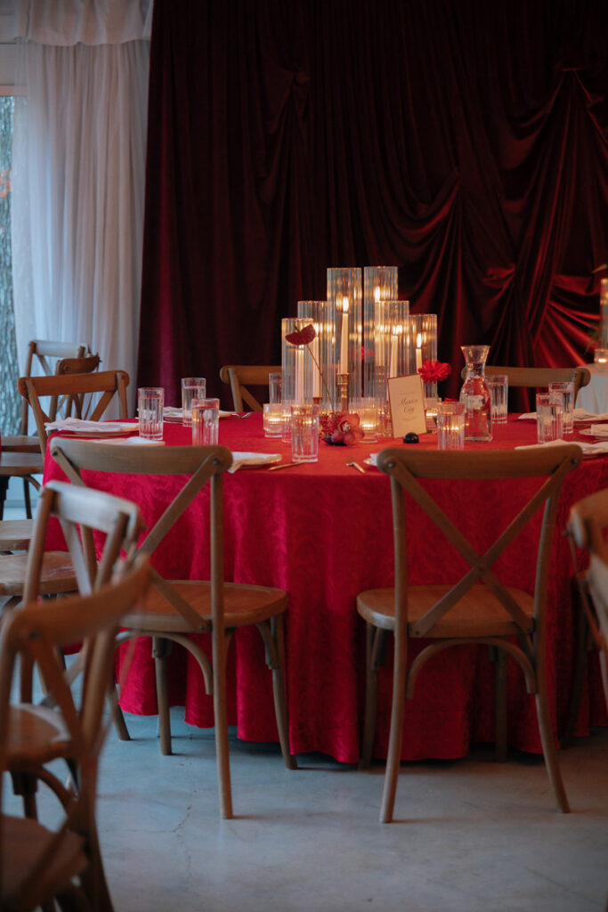 A round table covered with a red tablecloth, set with glassware, plates, and cutlery, and decorated with tall glass candle holders and small red flowers. Wooden chairs surround the table.