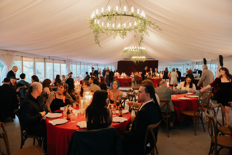 Guests seated at round tables with red tablecloths in a large, elegantly decorated wedding design ideas for tented wedding with chandeliers and draped white ceiling, attending a formal event or reception.