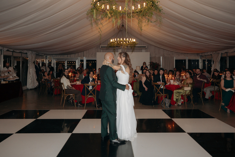 A bride and groom share their first dance on a black-and-white checkered floor in a large, elegant hall surrounded by seated guests.