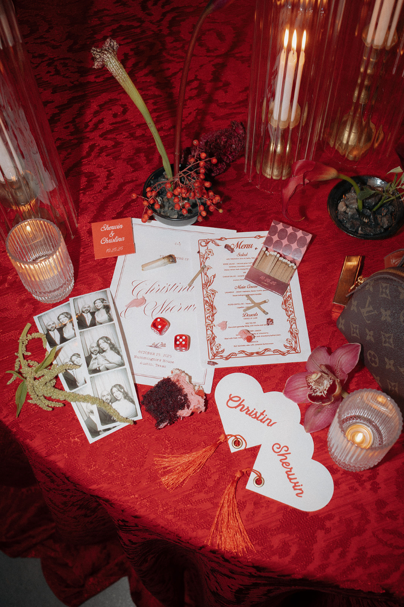 A red table holds candles, floral arrangements, personalized menus, a Louis Vuitton bag, photo booth strips, playing cards, dice, and heart-shaped name cards, perfect speakeasy wedding design ideas.