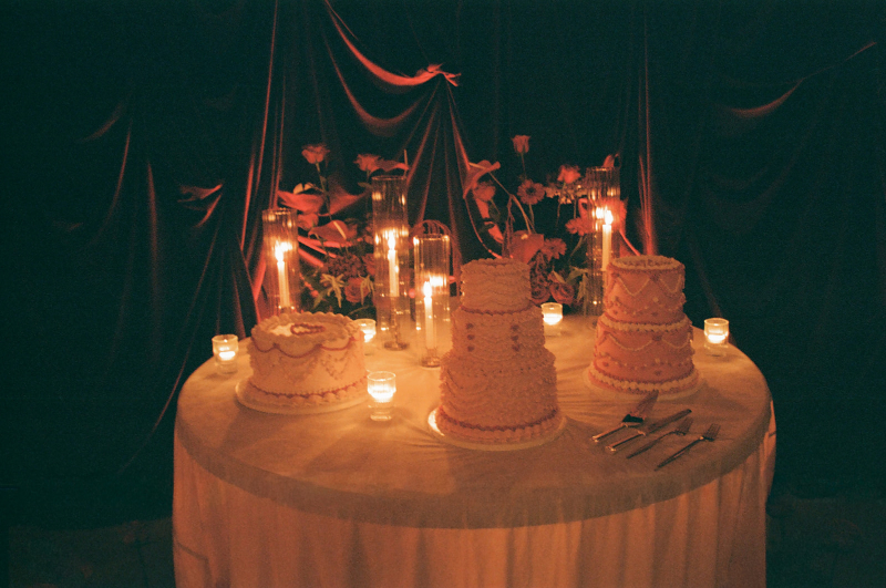 A round table with four decorated cakes, floral arrangements, and lit candles against a backdrop of draped dark fabric.