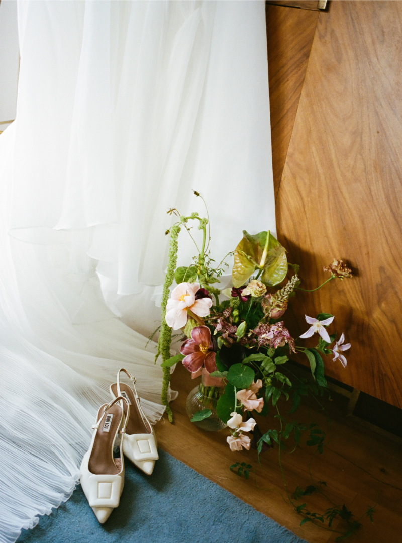 A pair of white heeled shoes, a bouquet of assorted flowers in a glass vase, and a white pleated dress draped beside a wooden surface—perfect inspiration for your $300k wedding budget checklist.