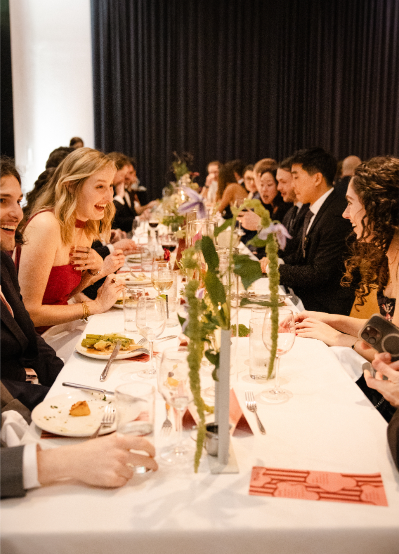 A group of people dressed in formal attire are seated at a long, decorated table, enjoying food and drinks, and conversing at an indoor celebration, possibly following a wedding budget checklist.