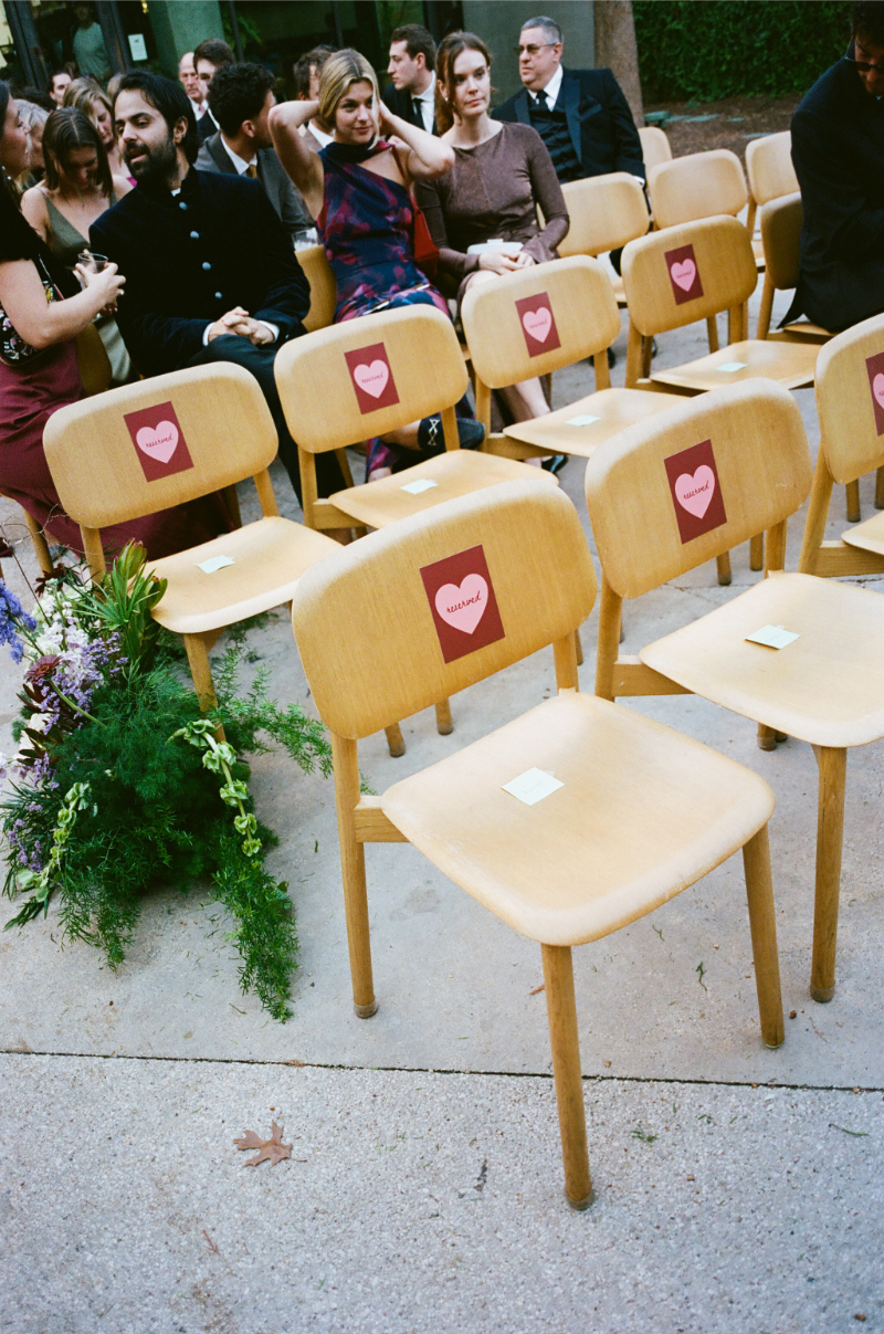 Several wooden chairs with heart-shaped reserved signs are arranged outdoors; a group of people sit and talk in the background, while flowers placed near the front row hint at thoughtful touches from a well-planned wedding budget checklist.
