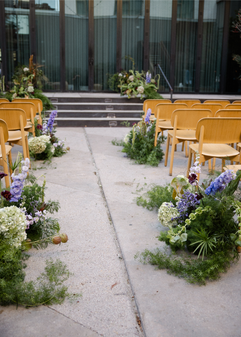 Rows of wooden chairs face a building with glass doors, flanked by lush floral arrangements along the aisle on a concrete surface—a perfect setting to consider as you finalize your wedding budget checklist.