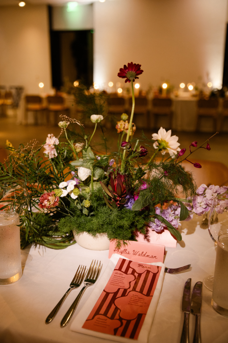 A table setting with a floral centerpiece, water glasses, cutlery, and a striped pink menu on a white tablecloth in a softly lit dining area—perfect inspiration for your wedding budget checklist.