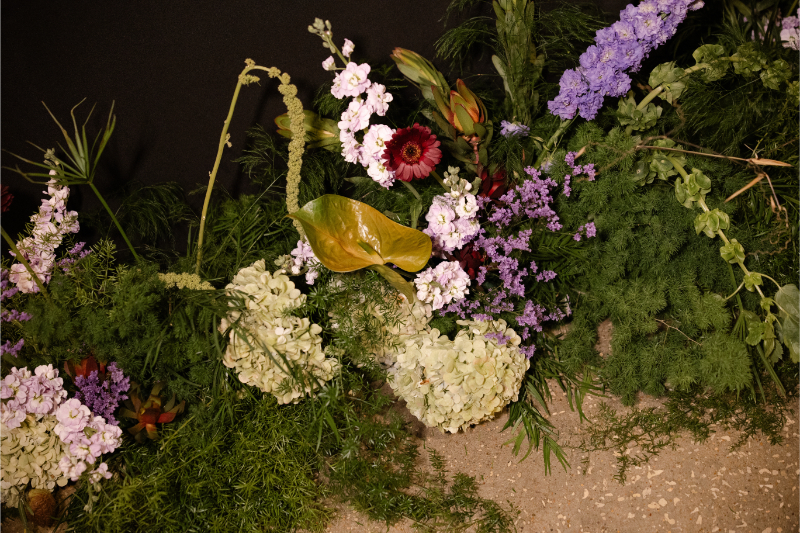 A variety of flowers and greenery, including hydrangeas, lilies, and purple blossoms, arranged against a dark background with soil at the bottom—perfect inspiration for your wedding budget checklist.
