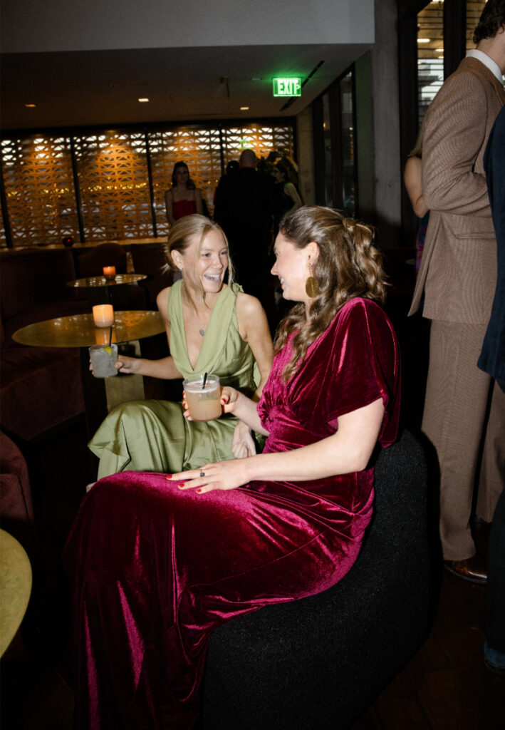 Two women in formal dresses sit and talk at a dimly lit lounge, each holding a drink. In the background, others mingle—perhaps discussing details like the wedding budget checklist for an upcoming celebration.