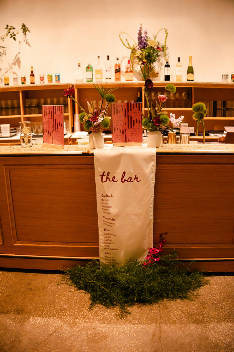A wooden bar counter with a drink menu sign reading "the bar," floral arrangements, and liquor bottles displayed on shelves in the background—a stylish detail to consider adding to your wedding budget checklist.