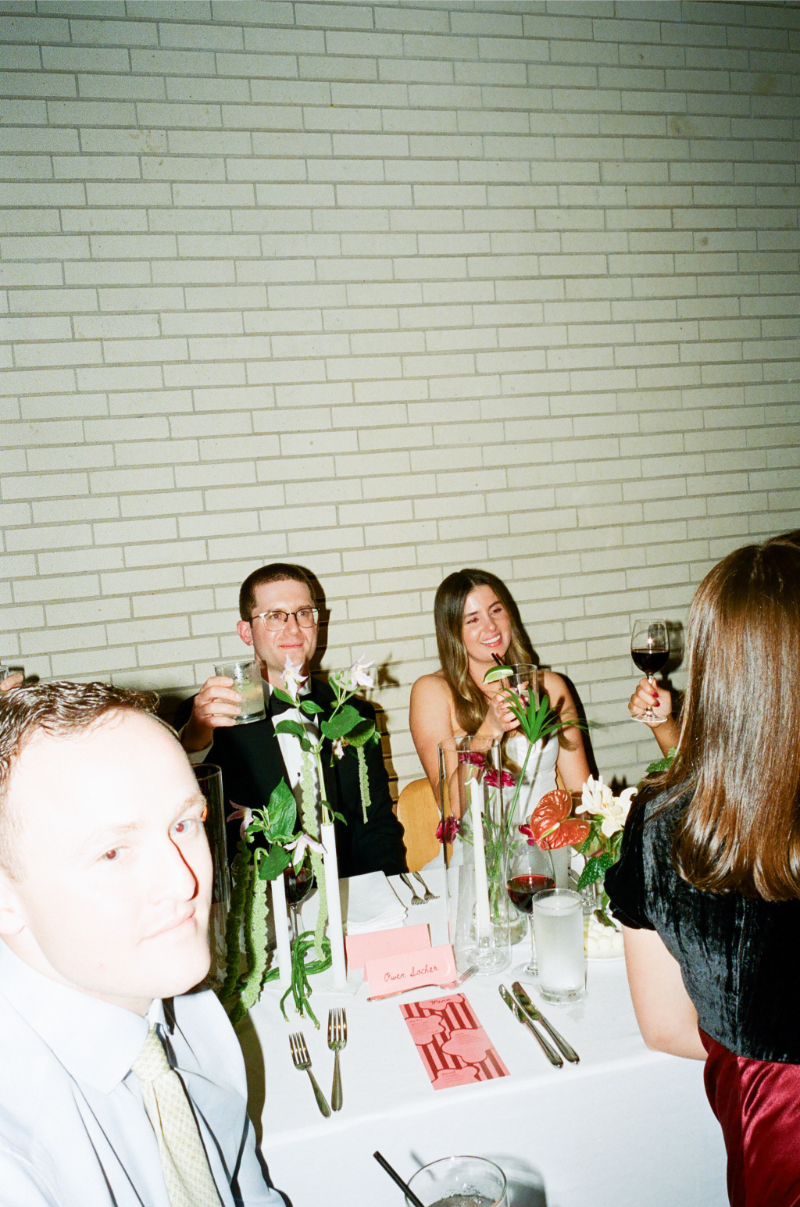 Four friends sit at a table set with drinks, flowers, a “Best Ladies” sign, and a wedding budget checklist, raising glasses in a toast against a white brick wall background.