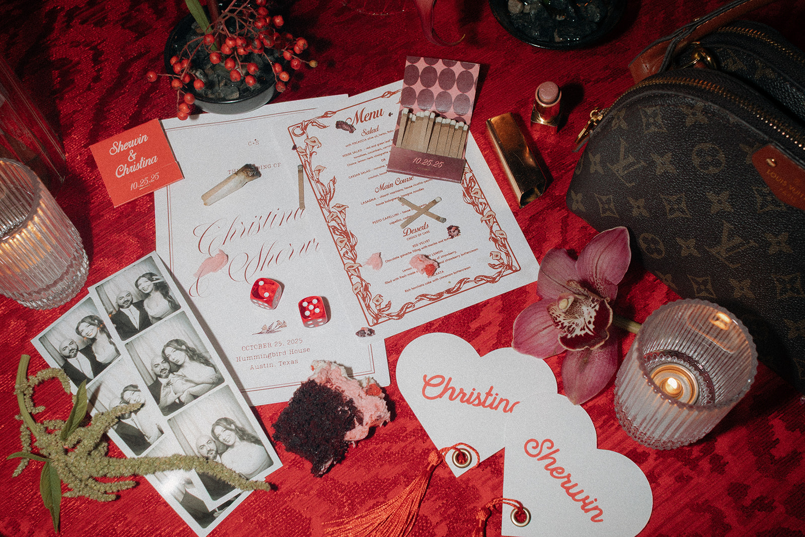 A flat lay of wedding invitations, menu, dice, photo booth strips, red velvet cake, lipstick, candles, and a Louis Vuitton bag arranged on a red surface with flowers.