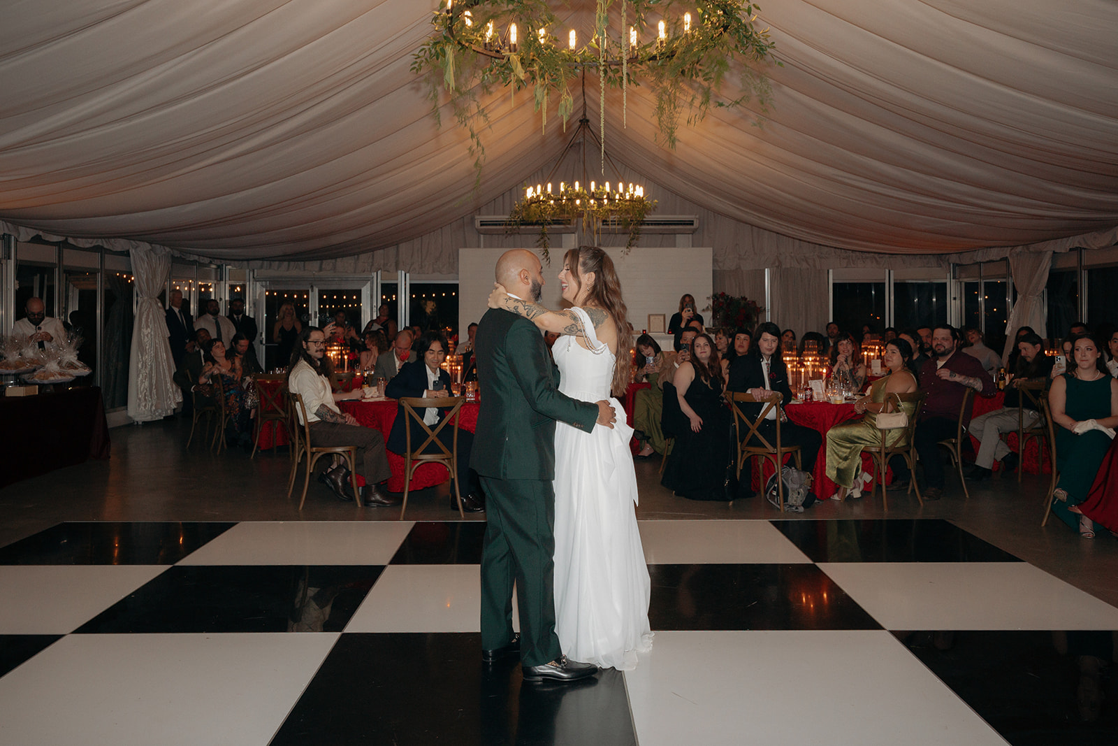 A bride and groom share their first dance in a decorated opera aesthetic wedding venue with guests seated at tables, watching them on a black-and-white checkered dance floor.