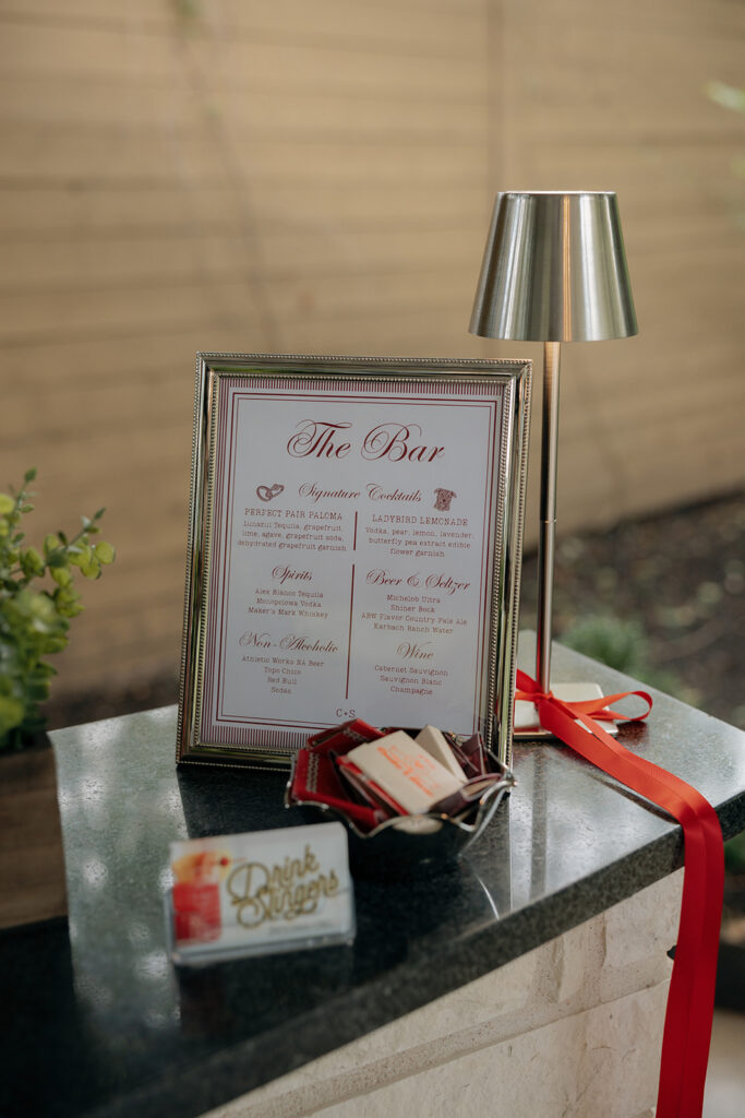 A framed bar menu with drink options stands on a countertop beside a small lamp, a red ribbon, and a card reading "Drink Signage" with wrapped mints.