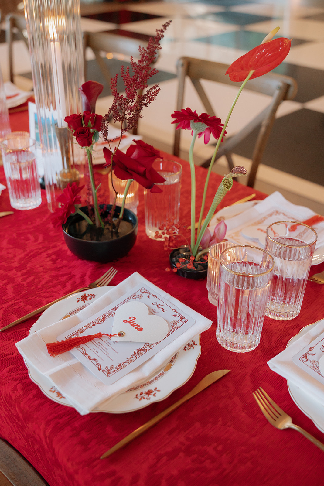 Elegant dinner table with red floral centerpiece, gold cutlery, clear glasses, and a menu card on a white plate atop a red tablecloth made for the perfect opera aesthetic wedding decor.
