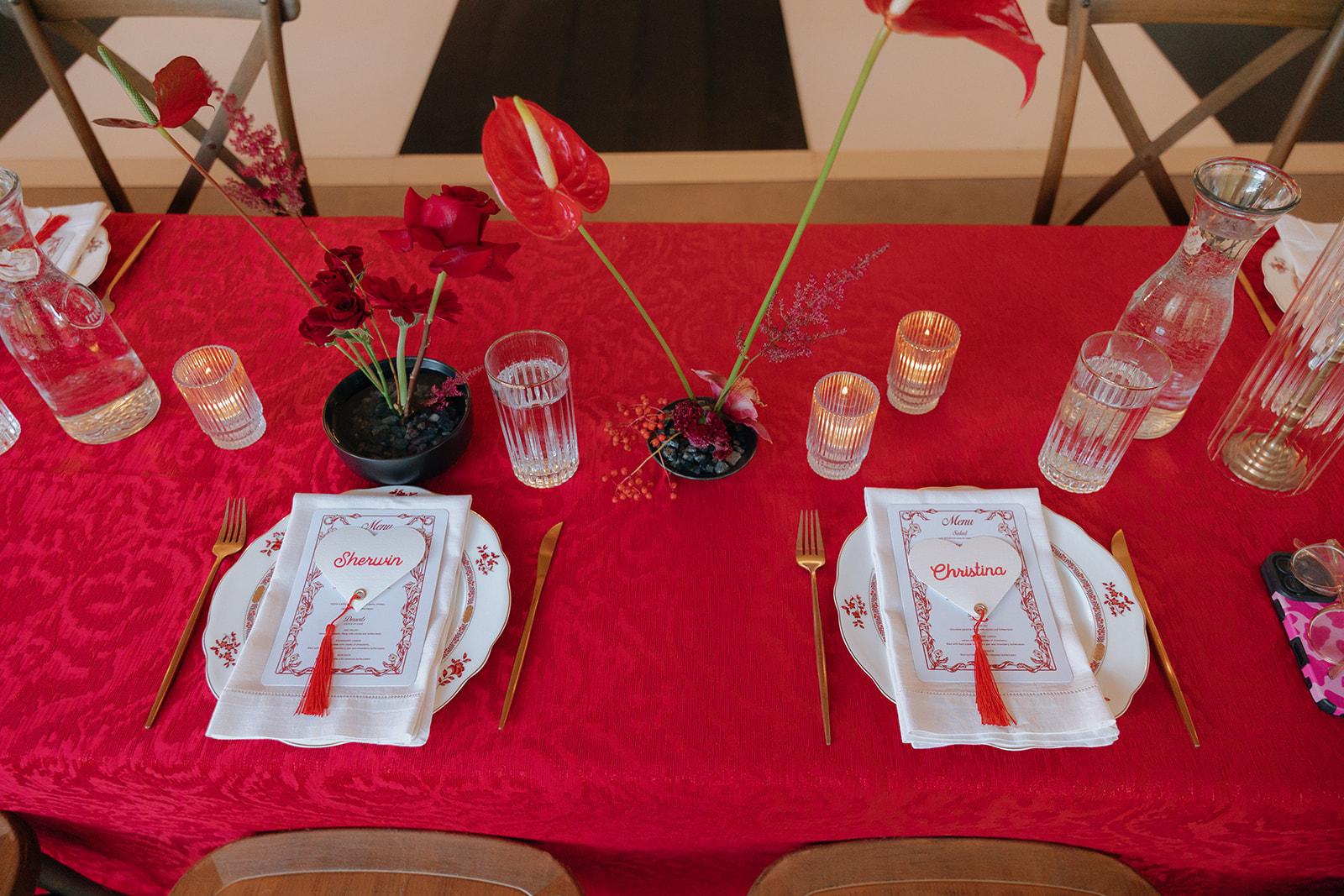 A red tablecloth with two place settings, gold cutlery, clear glassware, red floral arrangements, and candles on an opera aesthetic wedding tablescape for head table.