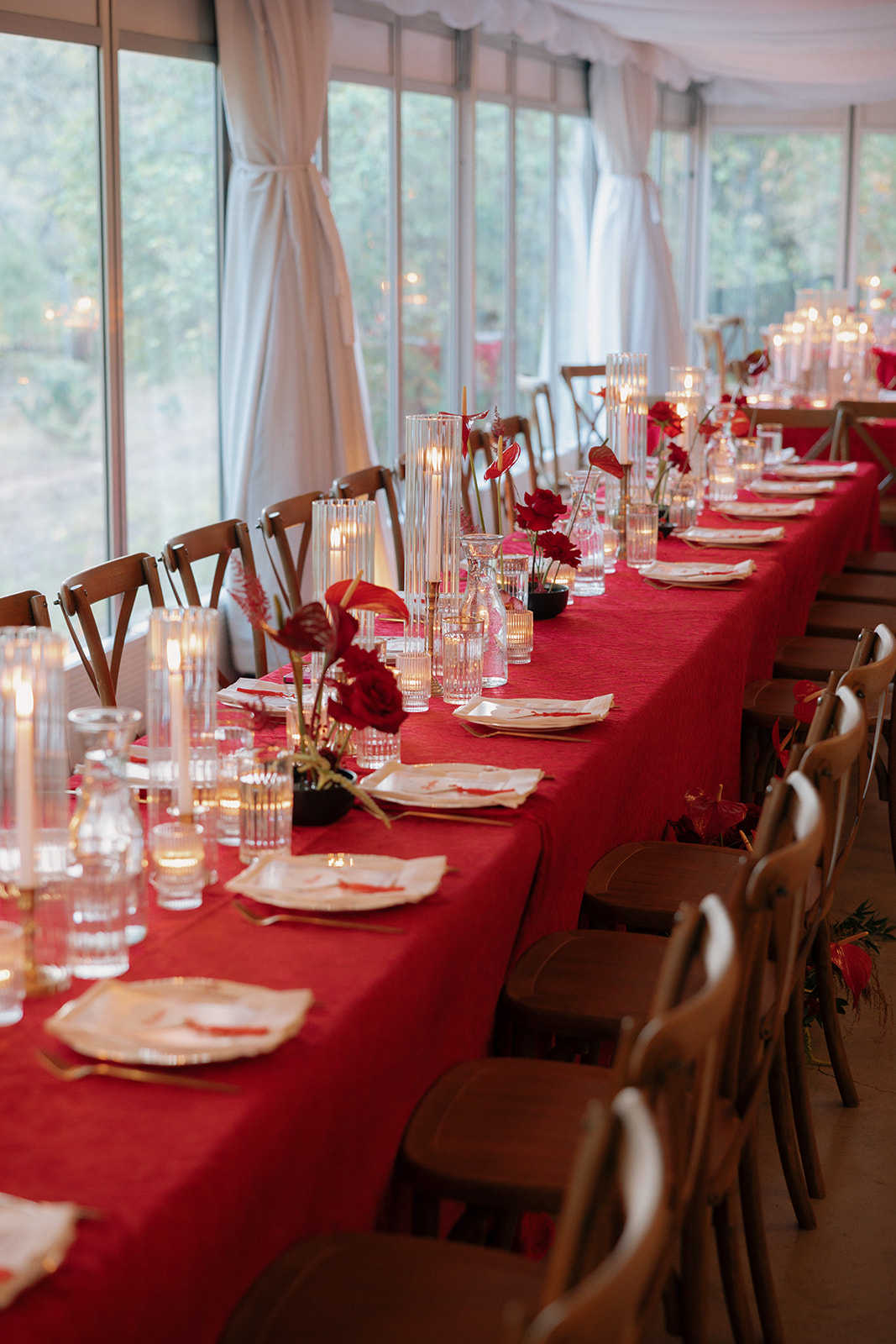 A long table with a red tablecloth is set with plates, glassware, tall glass candles, and small red floral arrangements in a room with large windows and white curtains for an opera aesthetic wedding.