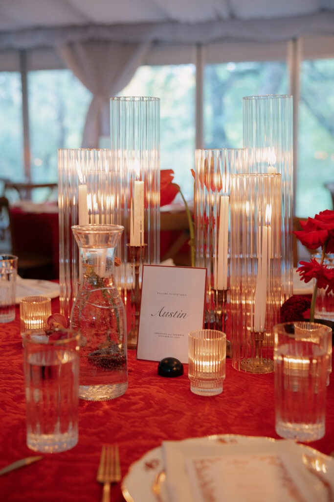 A formal table setting with ribbed glass candle holders, a glass water carafe, drinking glasses, red flowers, and a place card reading "Austin" on a red tablecloth.
