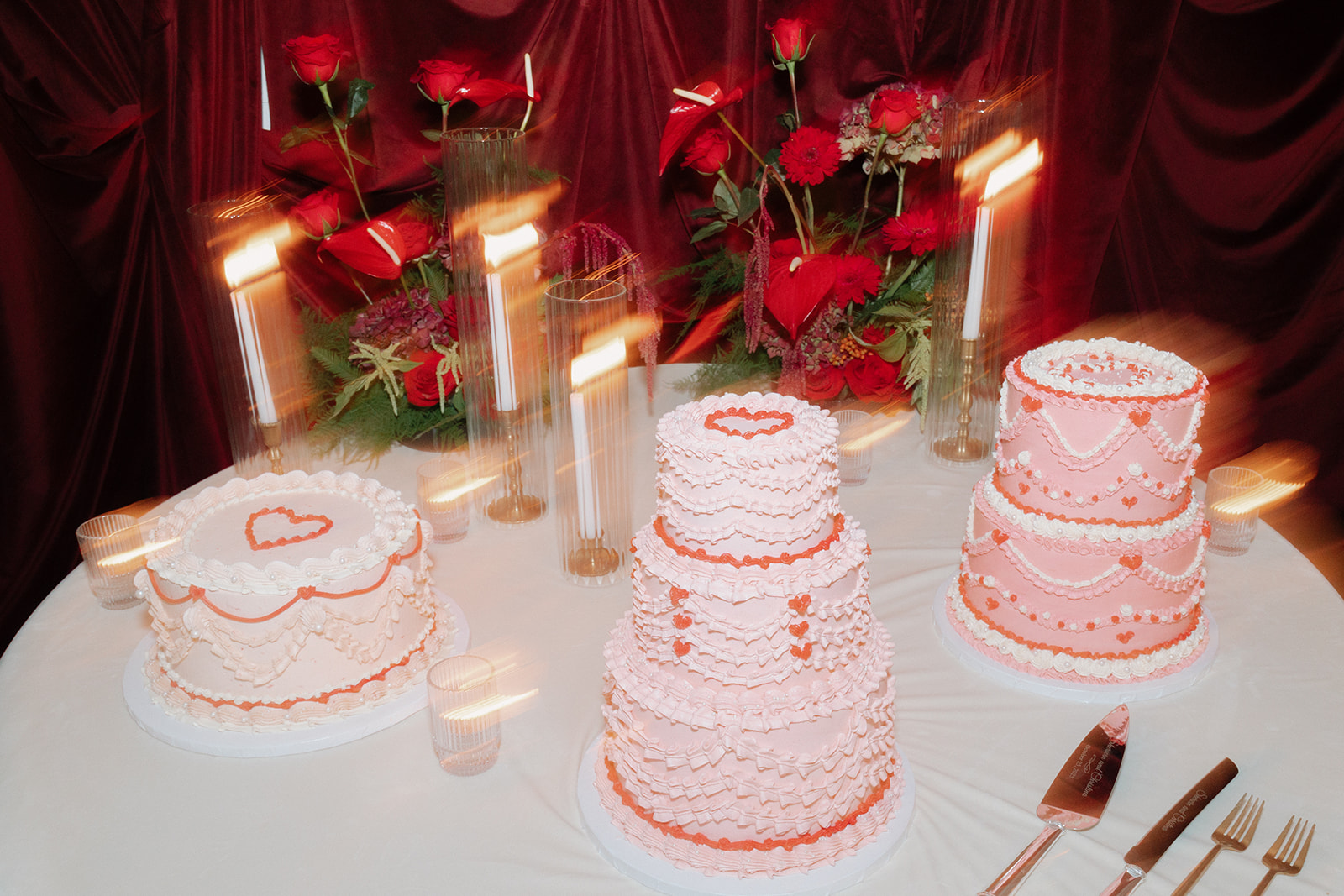 Three pink and white heart-themed cakes sit on a round table with utensils, surrounded by lit candles, red roses, and a maroon draped backdrop for an opera aesthetic wedding.