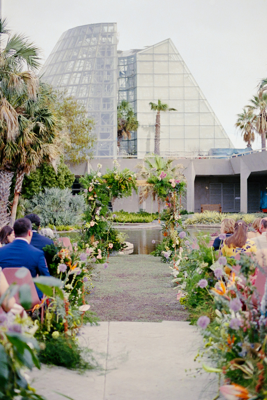 An outdoor wedding ceremony setup with floral arrangements along the aisle, guests seated, and glass pyramids and palm trees in the background.
