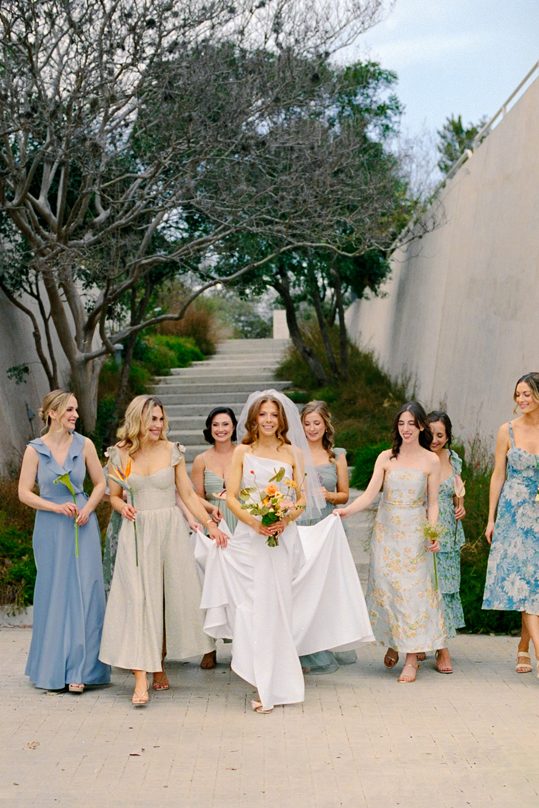 A bride in a white dress walks outdoors with six bridesmaids in pastel dresses, each holding a flower. Trees and concrete walls frame the background.