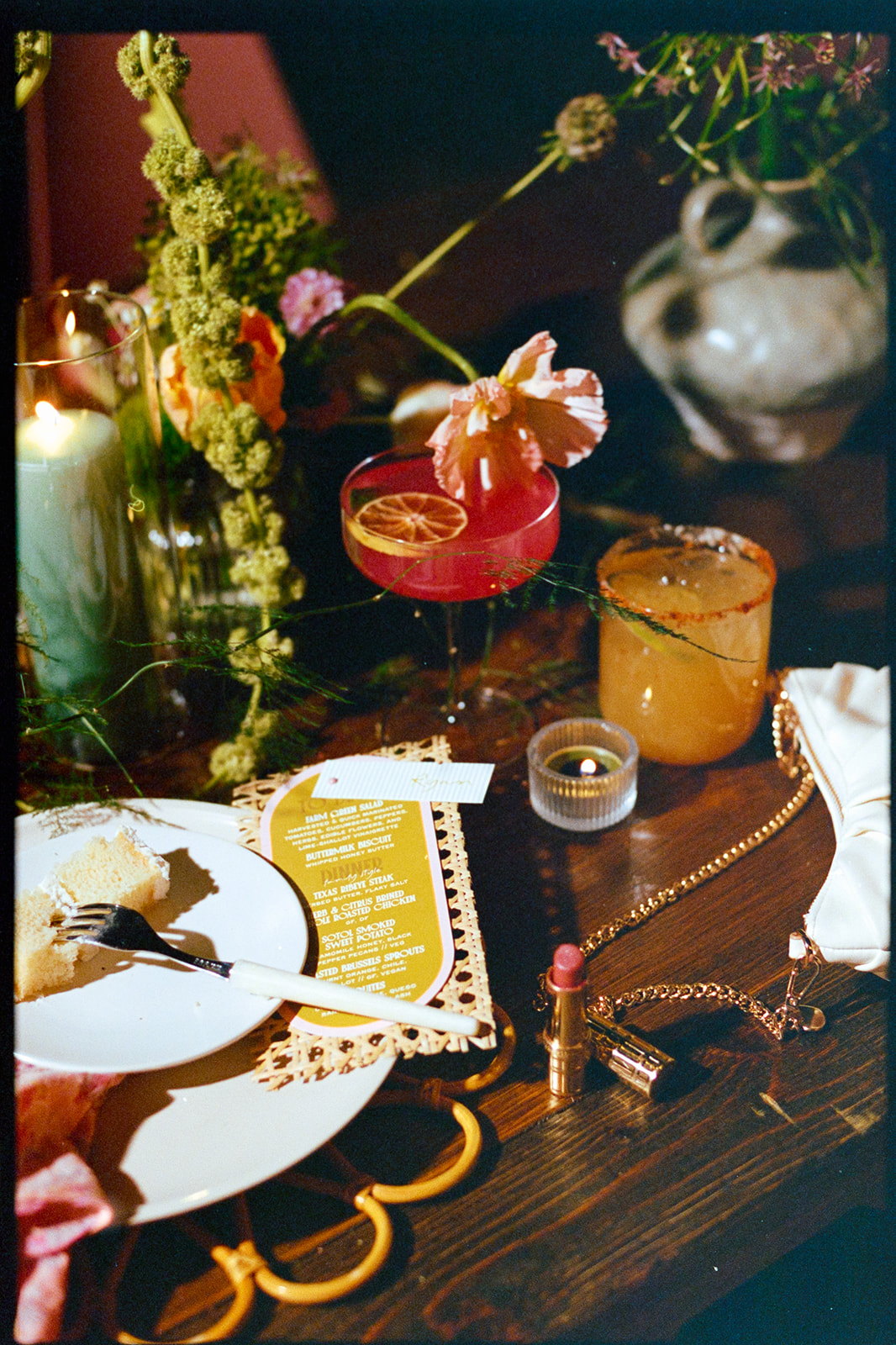 A table set with a partially eaten dessert, two cocktails, a lit candle, a lipstick, a purse, a menu, and floral decorations.