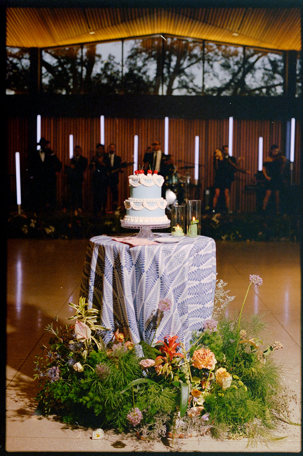 A tiered white cake with pink accents sits on a round, blue-patterned tablecloth surrounded by flowers; a band performs in the background.