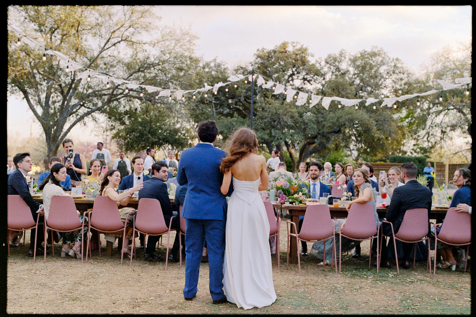 A couple stands arm in arm facing a long outdoor dining table filled with guests at a formal event, with string decorations overhead, the day designed by In Ink Weddings, an Austin and Santa Barbara wedding planner.