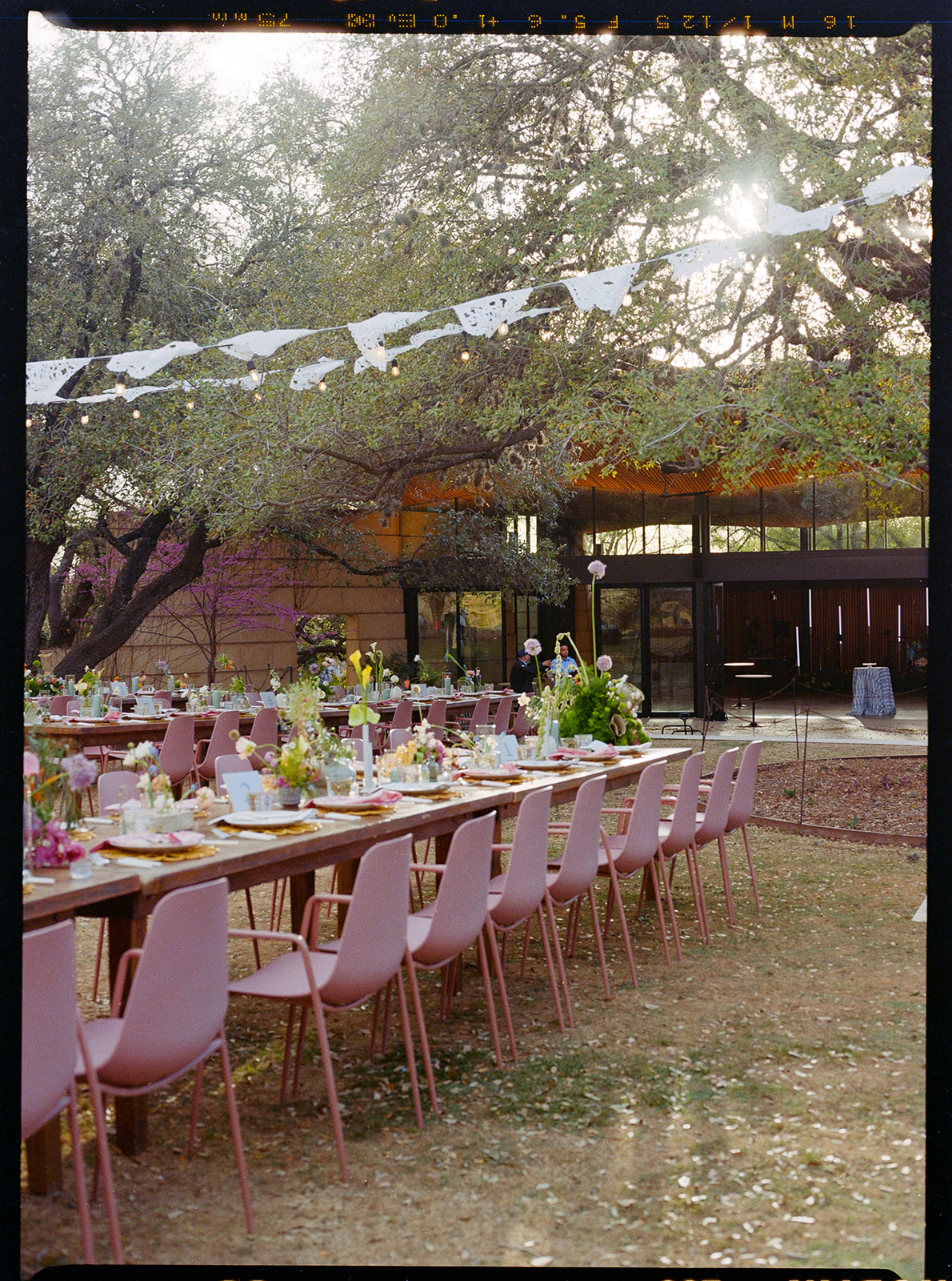 Long tables with pink chairs are set for an outdoor event under trees, with sunlight filtering through branches and white paper banners strung overhead, designed by In Ink Weddings, an Austin and Santa Barbara wedding planner.