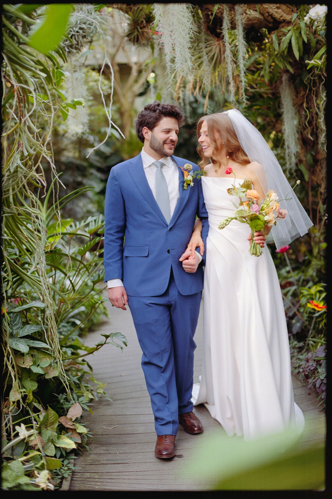 A bride in a white gown and veil holds a bouquet while walking hand in hand with a groom in a blue suit along a lush, plant-filled garden path.