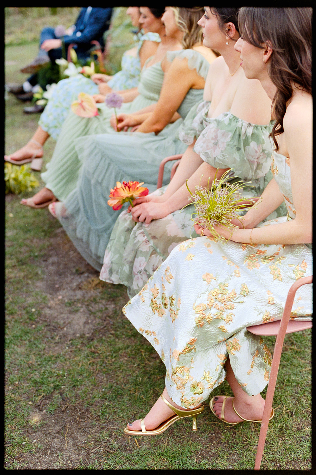 Women in pastel floral dresses sit on chairs outdoors, each holding a bouquet of flowers, with grass and more seated people visible in the background.