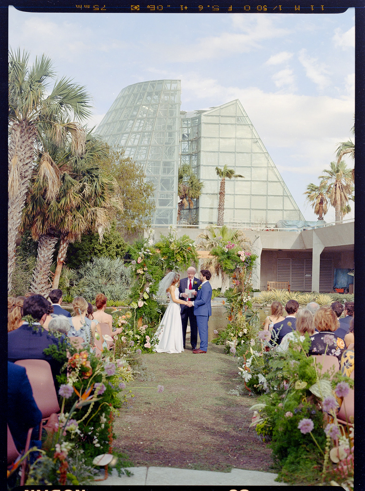 A bride and groom stand with an officiant in an outdoor wedding ceremony, surrounded by guests and greenery, with a glass conservatory in the background, designed by In Ink Weddings, an Austin and Santa Barbara wedding planner.