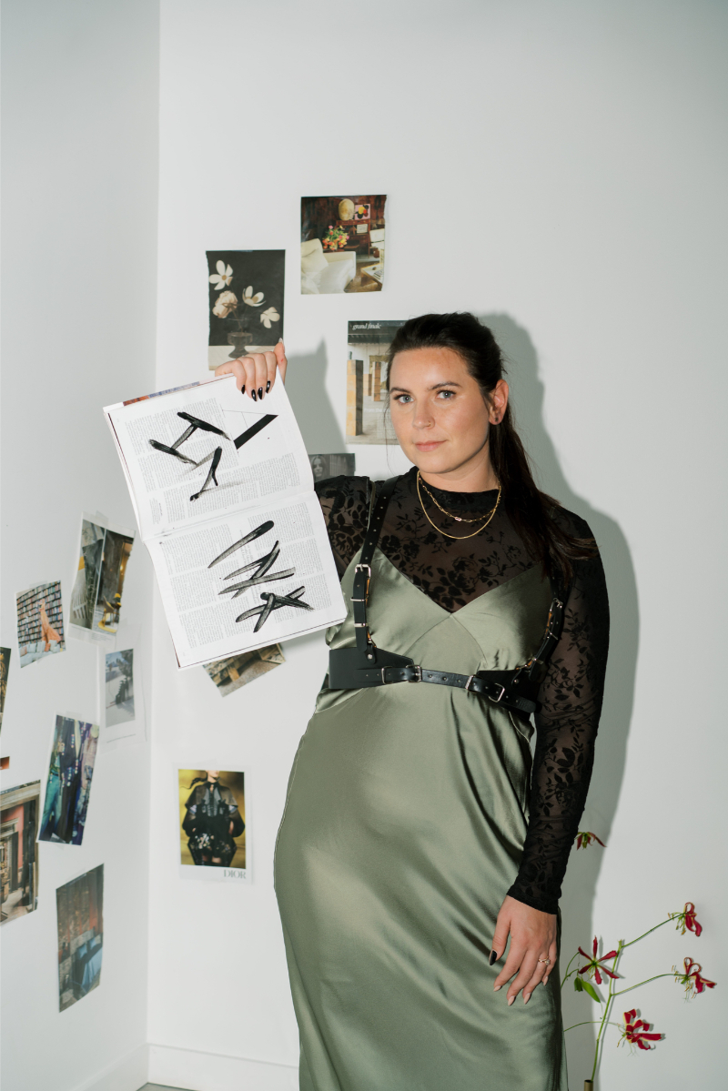 Syd from In Ink Weddings in a green satin dress and black lace top stands in a corner with art on the walls, holding up an open magazine behind the brand, displaying images of details behind the brand.