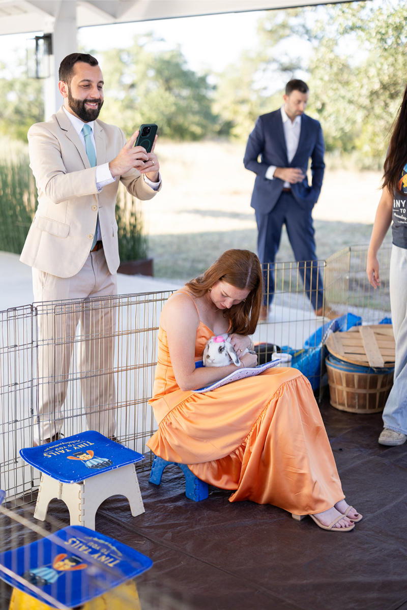 A woman in an orange dress sits inside a pen holding a white rabbit, while a man in a beige suit takes a photo with his phone. Two other people stand nearby.
