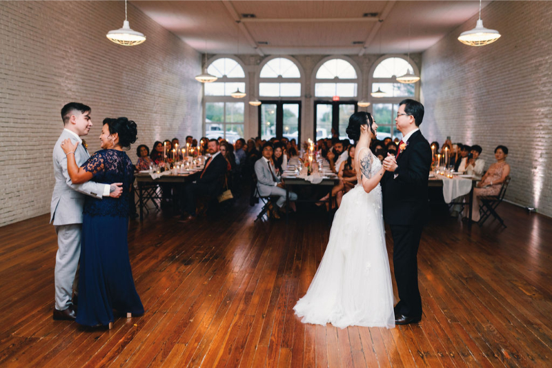 Two couples dance at a wedding reception in a hall with wooden floors and white brick walls, while guests seated at tables watch.