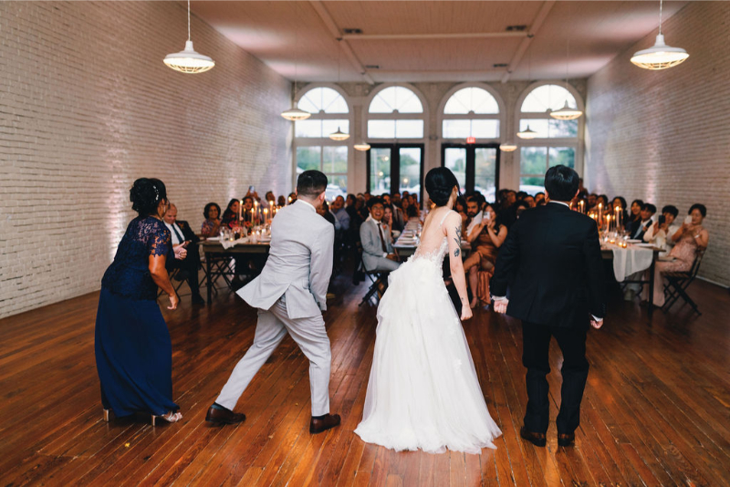 Four people, including a bride and groom, dance together on a wooden floor in front of seated wedding guests in a large, well-lit hall.