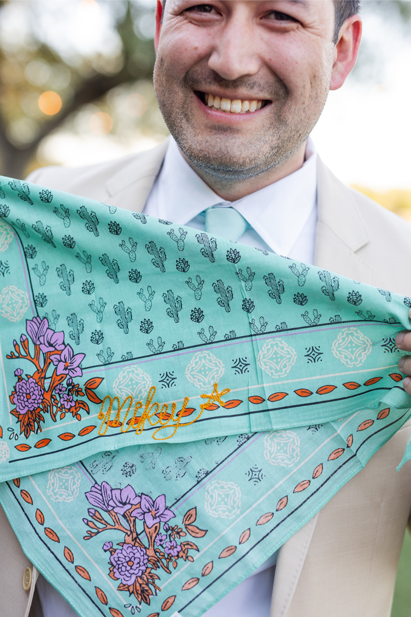 A man in a light suit holds a teal bandana with cactus and flower patterns and the name "Mikey's" embroidered in yellow thread.