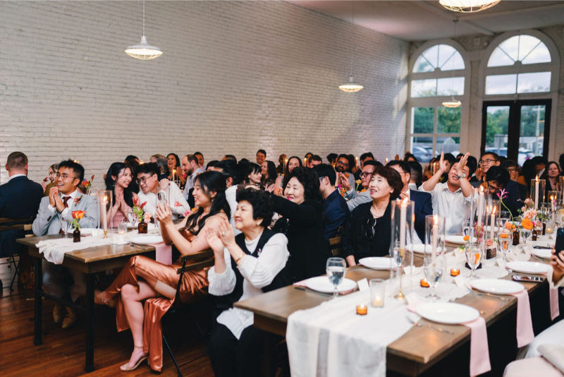 A group of people seated at decorated tables in a bright event space, clapping and smiling, likely attending a formal celebration or reception.